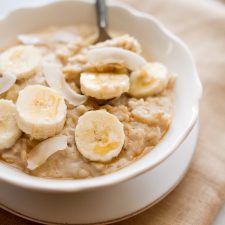 A white bowl of oatmeal covered with slices of banana, pieces of dried coconut, and a honey drizzle. A spoon is sitting in the bowl.