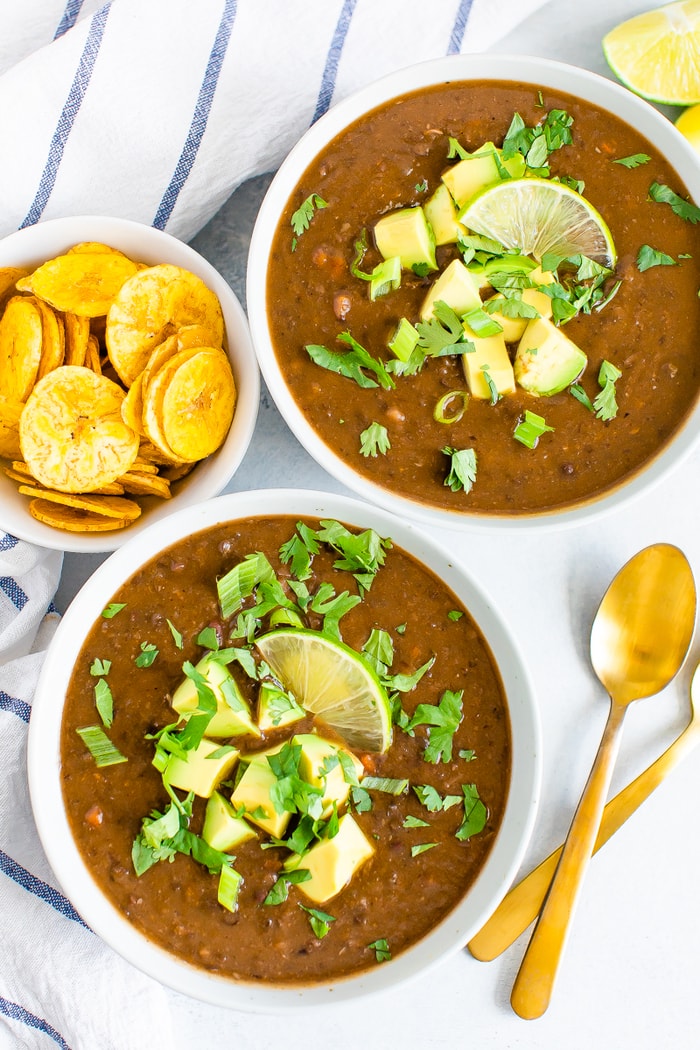 Two bowls of black bean soup topped with avocado, cilantro, green onion, and lime. Spoons, a dish cloth, and a bowl of plantain chips are next to the bowls.