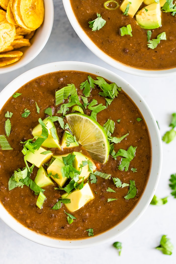 Two bowls of black bean soup topped with cilantro, green onion, avocado, and lime. A bowl of plantain chips are next to the bowls of black bean soup.