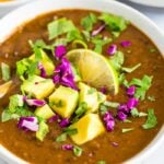 Bowl of black bean soup topped with avocado, cilantro, red cabbage, and lime.