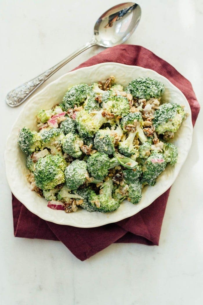 Ivory bowl with broccoli salad on red napkin. Silver spoon sitting next to bowl on marble countertop.