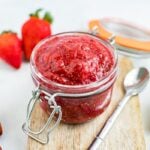 strawberry chia jam in a small open jar sitting on a wooden cutting board and surrounded by a few strawberries and a silver spoon.