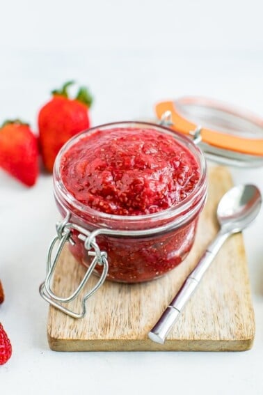 strawberry chia jam in a small open jar sitting on a wooden cutting board and surrounded by a few strawberries and a silver spoon.