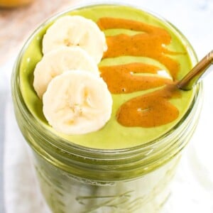Green smoothie in a mason jar, topped with banana slices and a peanut butter drizzle. A metal straw is in the jar. A bowl of peanut butter on a cutting board is out of focus in the background.