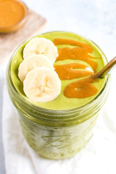 Green smoothie in a mason jar, topped with banana slices and a peanut butter drizzle. A metal straw is in the jar. A bowl of peanut butter on a cutting board is out of focus in the background.