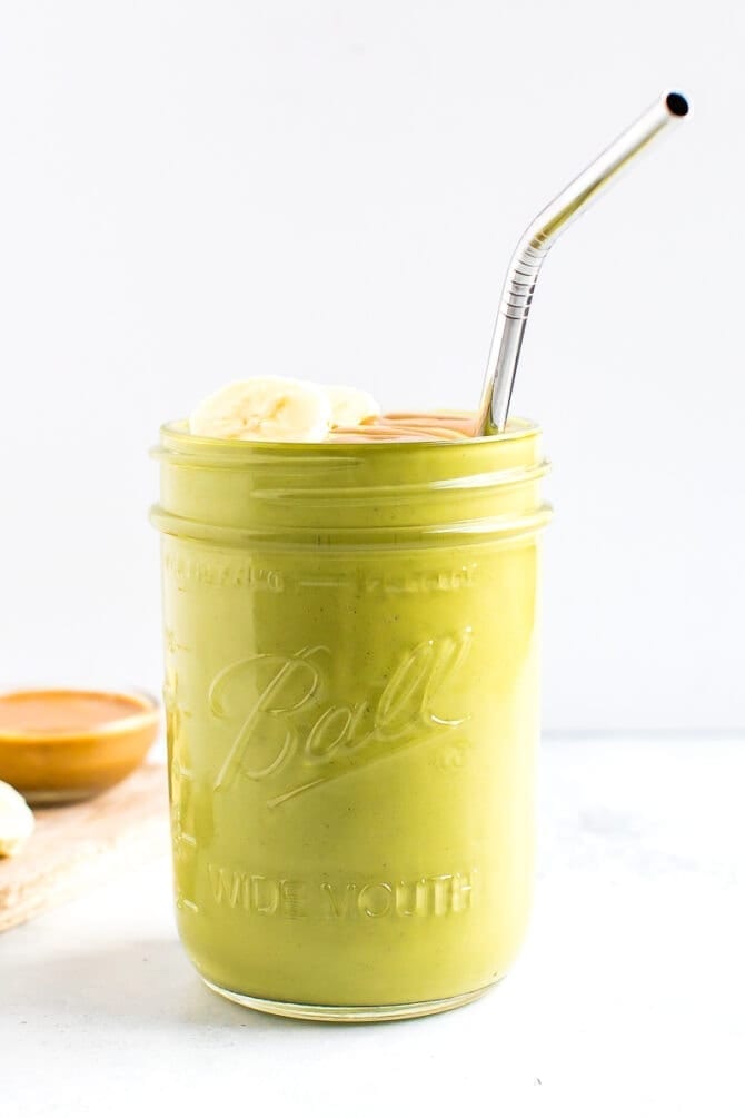 Green smoothie in a jar, topped with banana, peanut butter and a straw. A bowl of peanut butter is behind the jar.