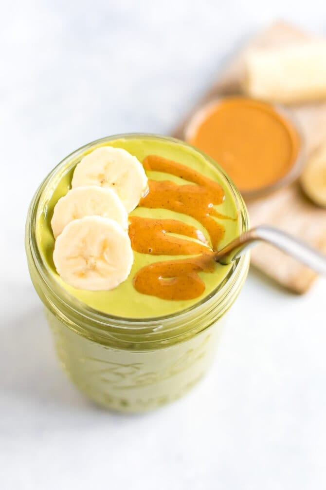 Green smoothie in a mason jar, topped with banana slices and a peanut butter drizzle. A metal straw is in the jar.
