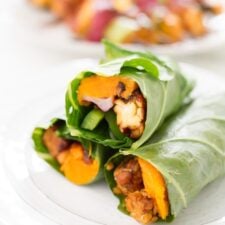 Three collard green wraps filled with, tempeh, creamy hummus, slices of baked sweet potato, green pepper, red onion and cranberries, stacked and sitting on a white plate.