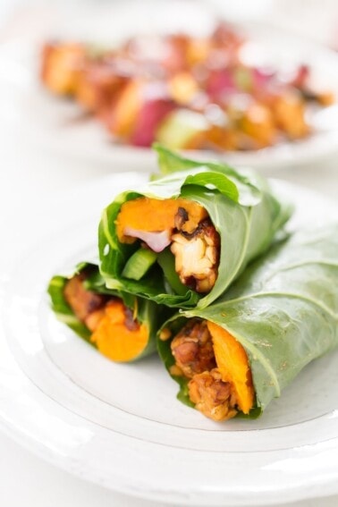 Three collard green wraps filled with, tempeh, creamy hummus, slices of baked sweet potato, green pepper, red onion and cranberries, stacked and sitting on a white plate.
