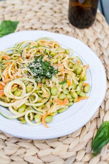 zucchini noodle pad Thai, with edamame, topped with chopped fresh basil leaves, on a white plate. A brown bottle is sitting in the background.