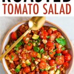 Serving bowl filled with a roasted tomato greek salad with a gold serving spoon resting on the bowl.