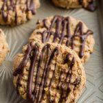 Four peanut butter granola cookies, drizzled with chocolate, sitting on a neutral colored tray.