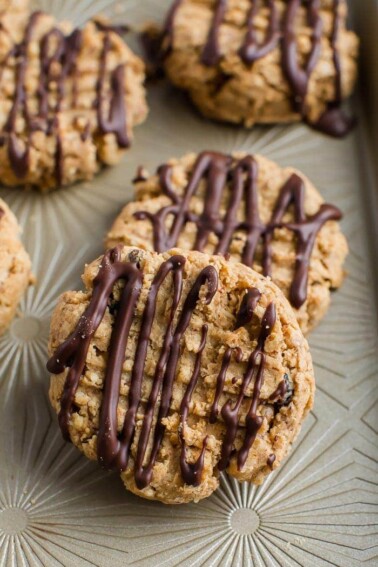 Four peanut butter granola cookies, drizzled with chocolate, sitting on a neutral colored tray.