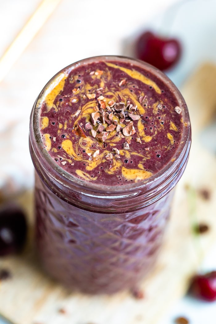 Mocha cherry smoothie in a mason jar topped with cacao nibs, and a swirl of almond butter. Cherries are on the board around the smoothie.