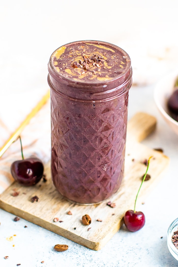 Mocha cherry smoothie in a mason jar topped with cacao nibs and almond butter swirl. Cherries and coffee beans are around the jar.