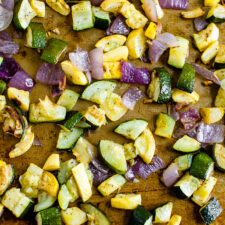 Roasted, chopped summer squash, zucchini, and red onion on a sheet with spices and pepper sprinkled on them.