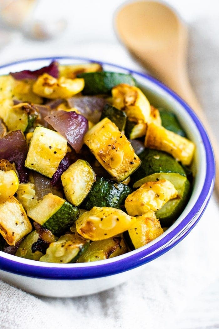 Bowl of roasted summer squash, zucchini, and red onion with pepper and seasoning. A wooden spoon is beside the bowl.