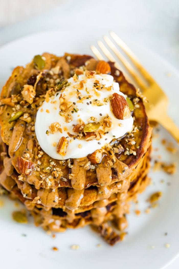 Stack of butternut squash pancakes topped with peanut butter, maple syrup, granola, and yogurt.