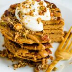 Stack of butternut squash pancakes topped with peanut butter, maple syrup, granola, and yogurt. A bite is cut out of the stack of pancakes and a gold fork is sitting next to the stack.