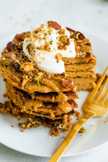 Stack of butternut squash pancakes topped with peanut butter, maple syrup, granola, and yogurt. A bite is cut out of the stack of pancakes and a gold fork is sitting next to the stack.