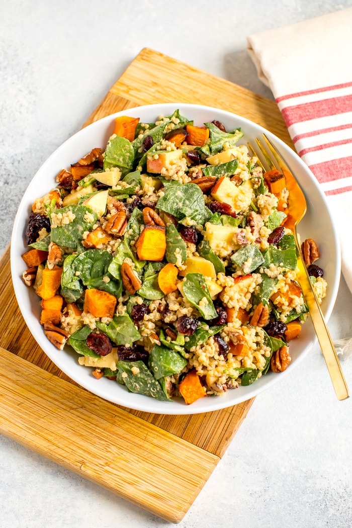 Salad plate filled with spinach harvest salad including butternut squash, cranberries, quinoa, pecans, and apples. A fork of on the plate and a dish towel to the side.
