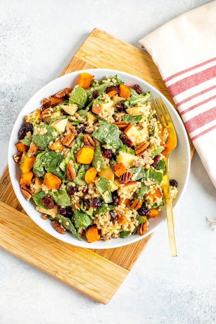 Salad plate filled with spinach harvest salad including butternut squash, cranberries, quinoa, pecans, and apples. A fork of on the plate and a dish towel to the side.