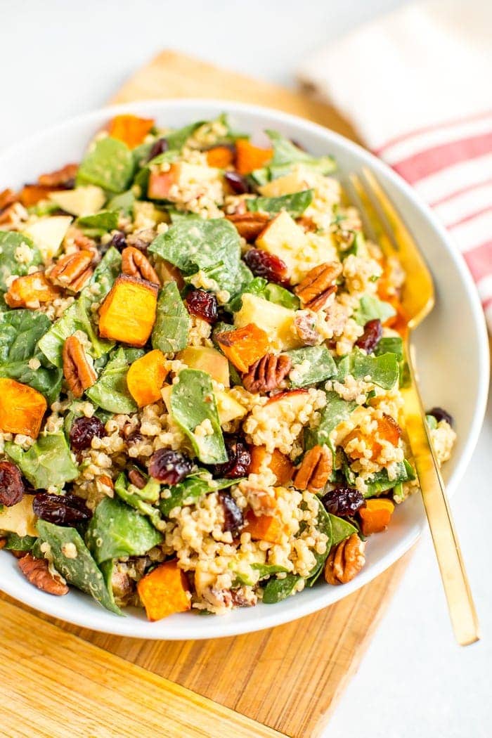 Salad plate filled with spinach harvest salad including butternut squash, cranberries, quinoa, pecans, and apples. A fork of on the plate and a dish towel to the side.