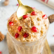Glass of healthy eggnog overnight oats topped with pomegranate seeds. A spoon is taking a spoonful of the creamy oats.