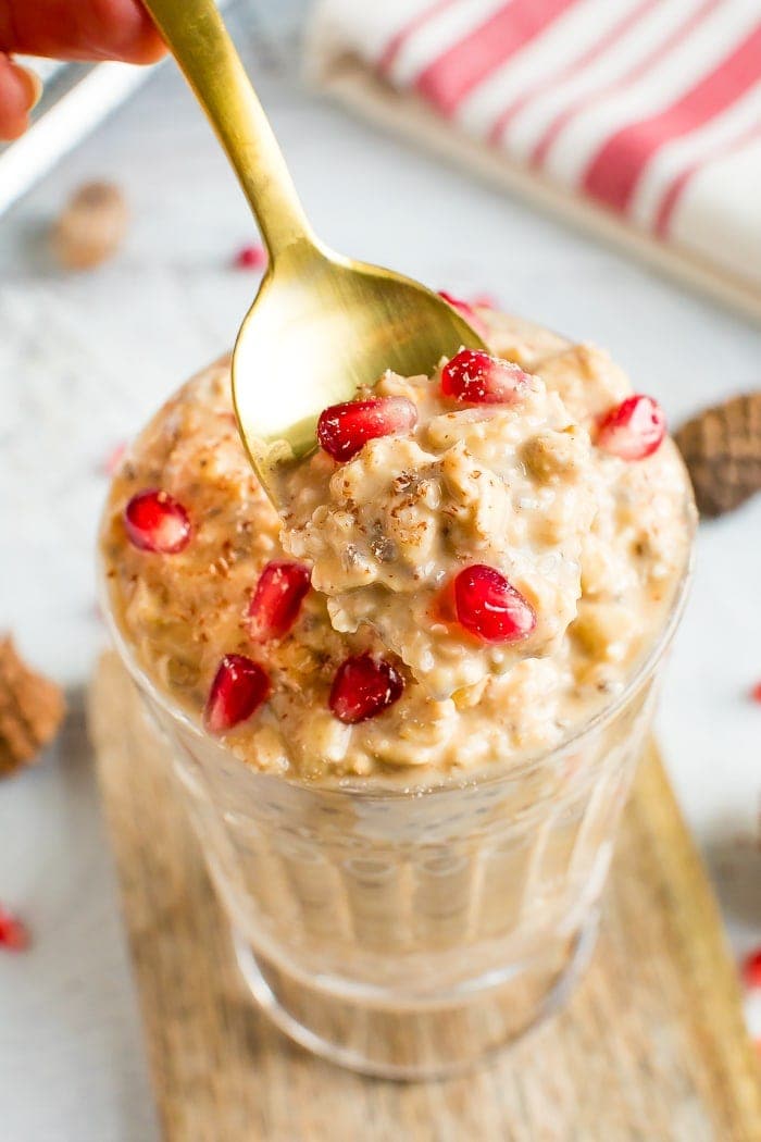 Glass of healthy eggnog overnight oats topped with pomegranate seeds. A spoon is taking a spoonful of the creamy oats.