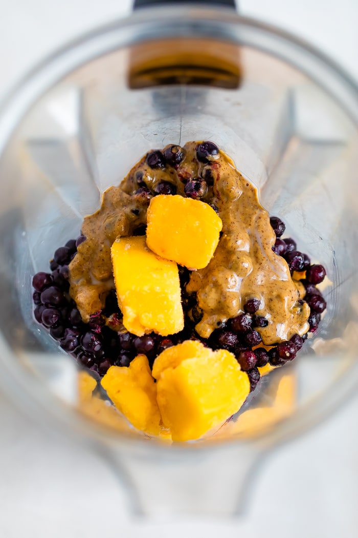 Ingredients for the health nut smoothie in the blender: frozen mango, frozen blueberries, frozen banana, protein powder, almond butter, and almond milk.