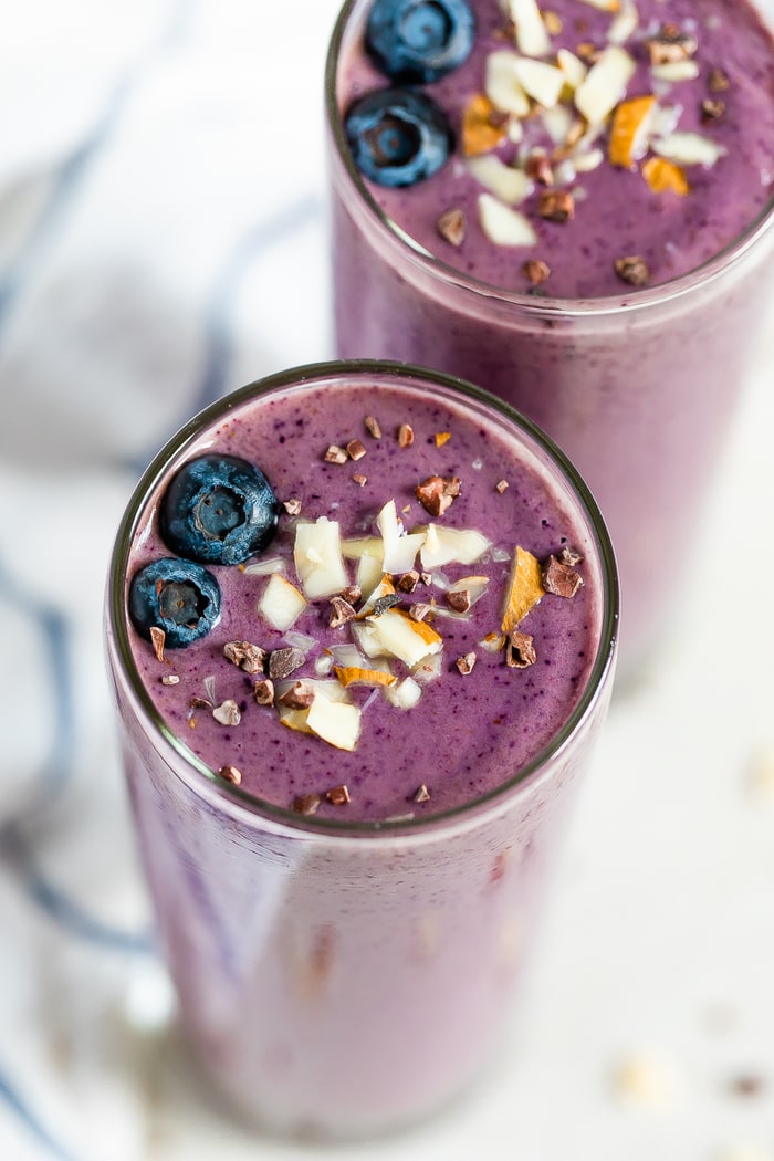 Two purple health nut smoothies topped with blueberries, chopped almonds, and cacao nibs.