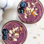 Two purple health nut smoothies topped with blueberries, chopped almonds, and cacao nibs.