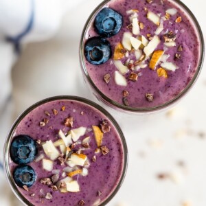 Two purple health nut smoothies topped with blueberries, chopped almonds, and cacao nibs.