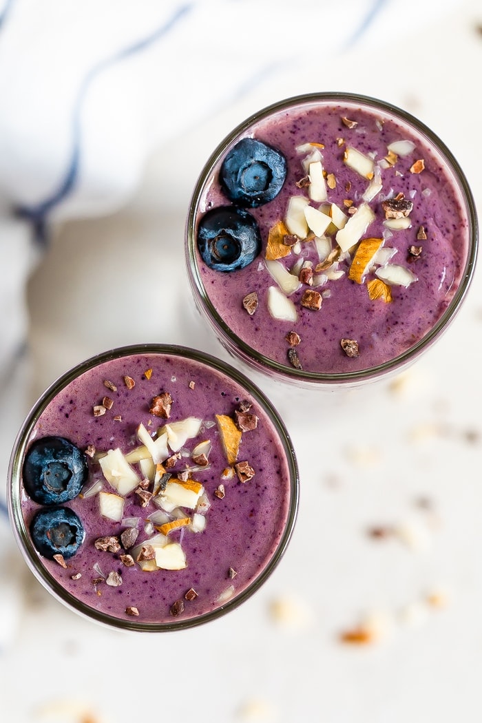Two purple health nut smoothies topped with blueberries, chopped almonds, and cacao nibs.