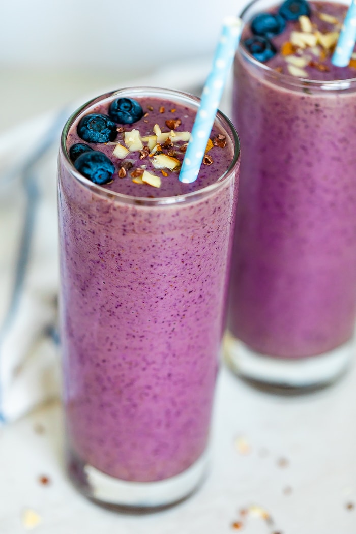 Two purple smoothies topped with chopped almonds, blueberries, cacao nibs, and with two paper polka dot blue straws.