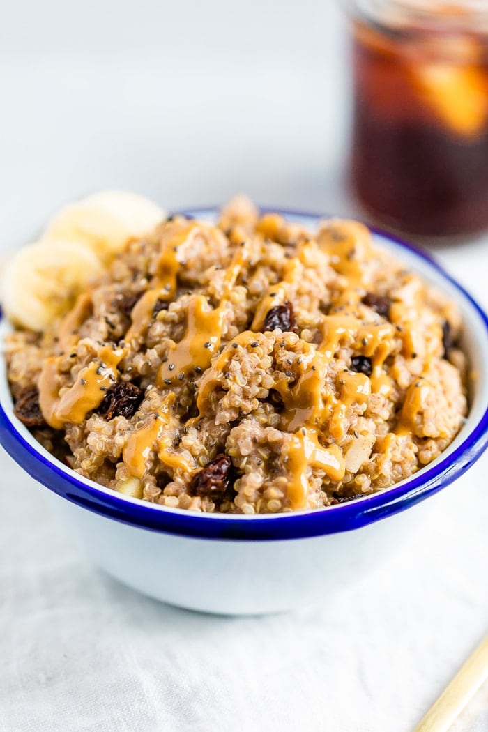 Bowl of cinnamon breakfast quinoa, topped with raisins, chia seeds, peanut butter, and banana slices. A glass of iced coffee is beside the bowl.