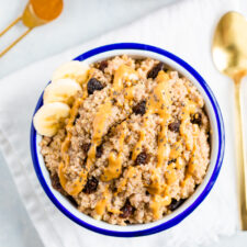 Bowl of cinnamon breakfast quinoa, topped with raisins, chia seeds, peanut butter, and banana slices. A teaspoon of cinnamon, a gold spoon, and a glass of iced coffee are beside the bowl.