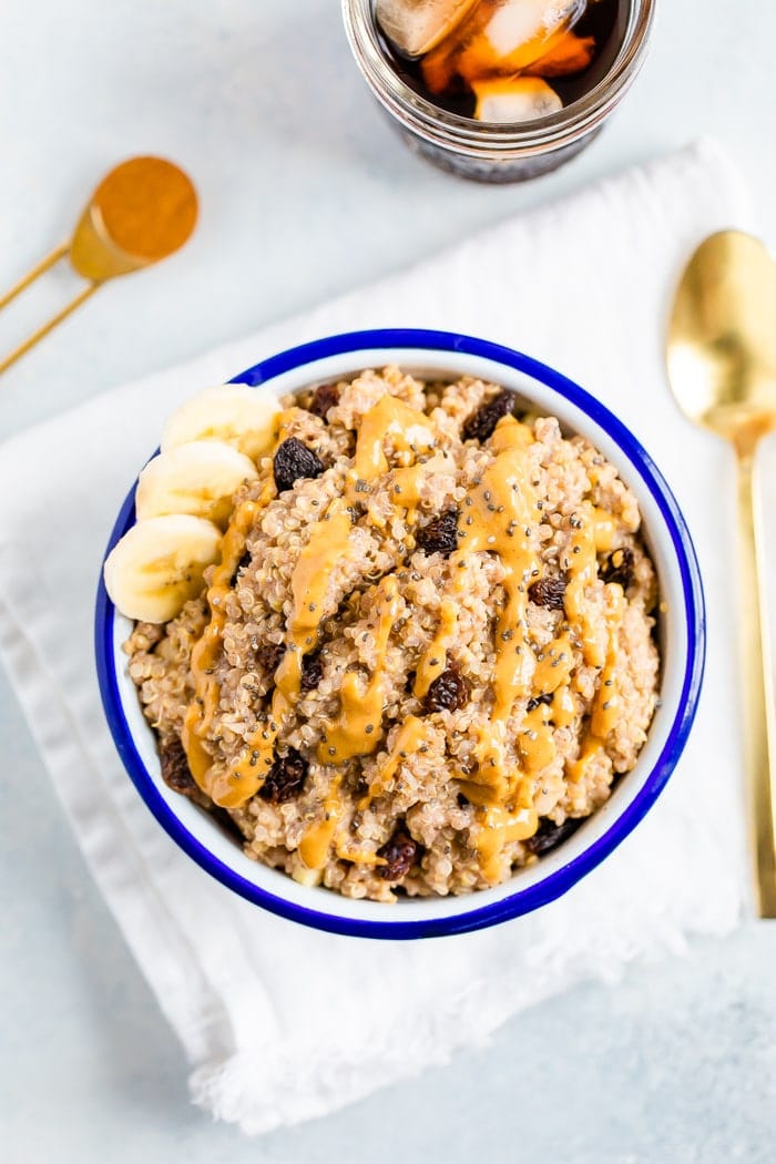 Bowl of cinnamon breakfast quinoa, topped with raisins, chia seeds, peanut butter, and banana slices. A teaspoon of cinnamon, a gold spoon, and a glass of iced coffee are beside the bowl.