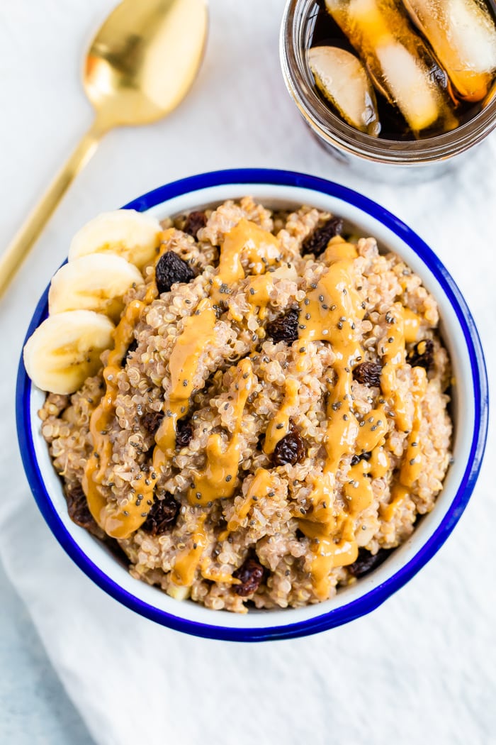 Bowl of cinnamon breakfast quinoa, topped with raisins, chia seeds, peanut butter, and banana slices. A gold spoon and jar of coffee are to the side.