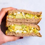 A hand holding curried avocado egg salad in a sandwich that is cut in half..