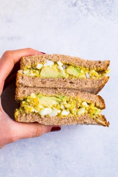 A hand holding curried avocado egg salad in a sandwich that is cut in half..