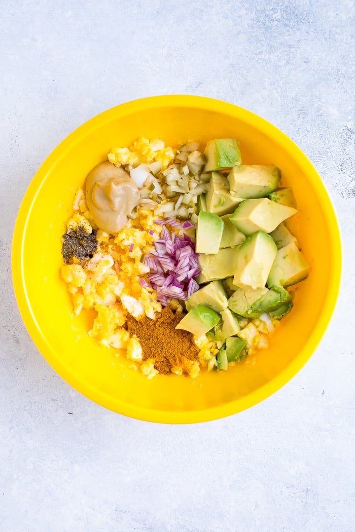 A yellow bowl with all the ingredients to make curried avocado egg salad.