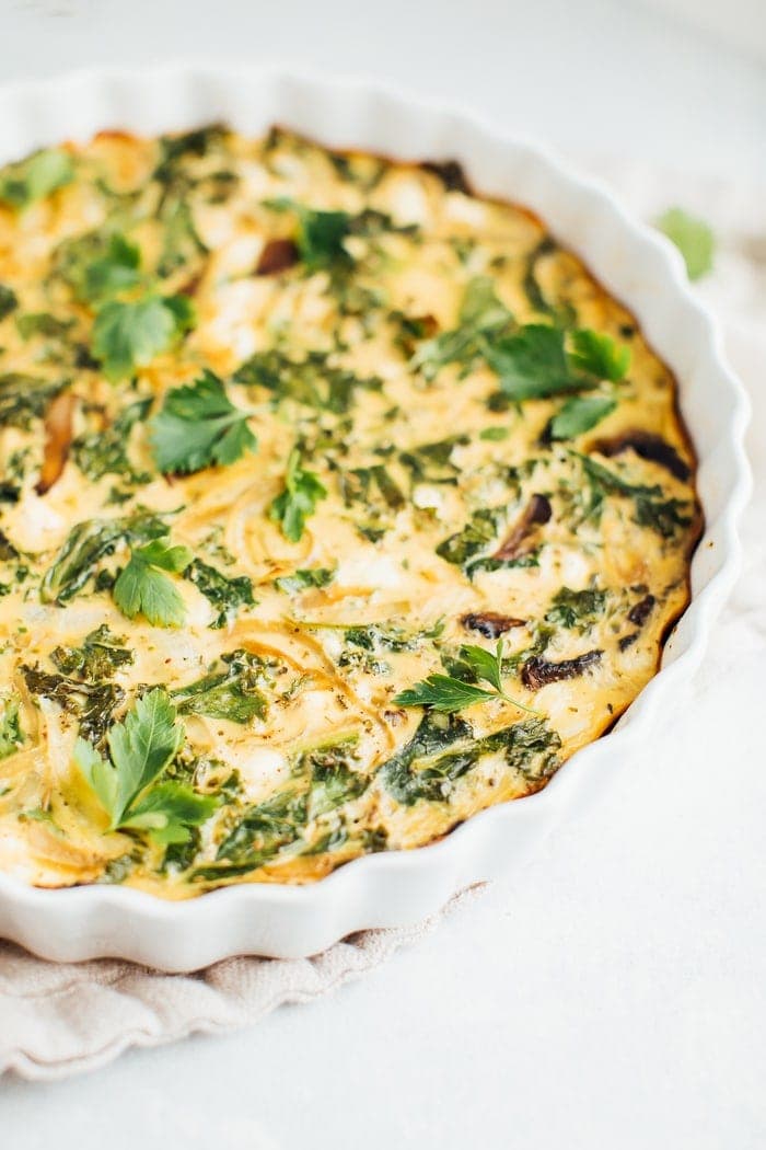 Crustless quiche with kale, onions, mushrooms and feta in a white baking dish on a rustic napkin.
