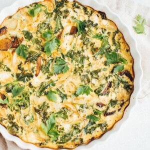 Crustless quiche with kale, onions, mushrooms and feta in a white baking dish on a rustic napkin.
