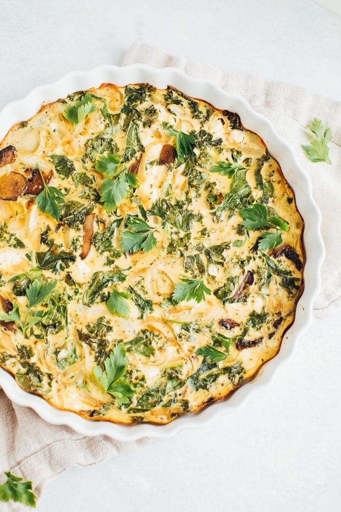 Crustless quiche with kale, onions, mushrooms and feta in a white baking dish on a rustic napkin.