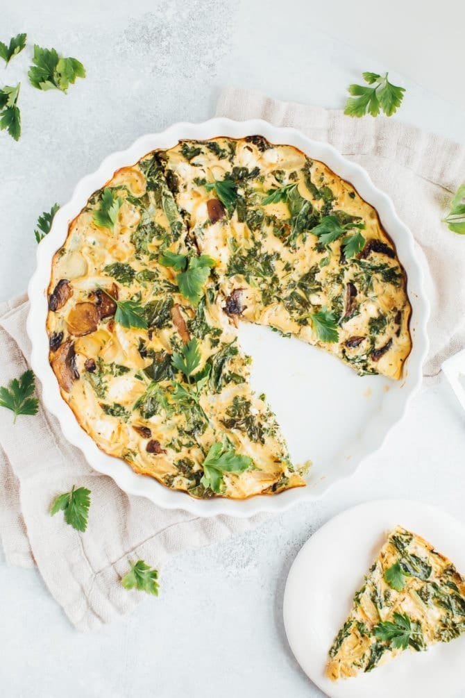 View from above of crustless quiche with kale, onions, mushrooms and feta in a white baking dish on a rustic napkin with fresh parsley around the dish for garnish. One slice is cut out of quiche and sitting on a separate white plate.
