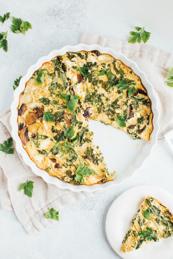 Crustless quiche with kale, onions, mushrooms and feta in a white baking dish on a rustic napkin with fresh parsley around the dish for garnish.