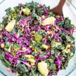 Salad with kale, cabbage, avocado, broccoli, chickpeas, and a nutritional yeast goddess dressing in a bowl. Wooden spoon sitting in the bowl too.