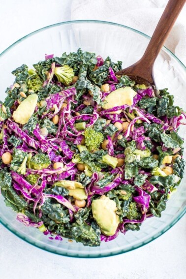 Salad with kale, cabbage, avocado, broccoli, chickpeas, and a nutritional yeast goddess dressing in a bowl. Wooden spoon sitting in the bowl too.