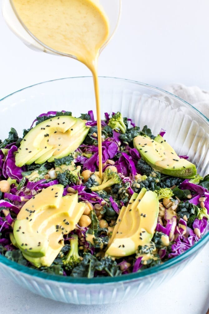 Nutritional yeast goddess dressing being poured over a kale and cabbage salad with broccoli, chickpeas, and avocado.